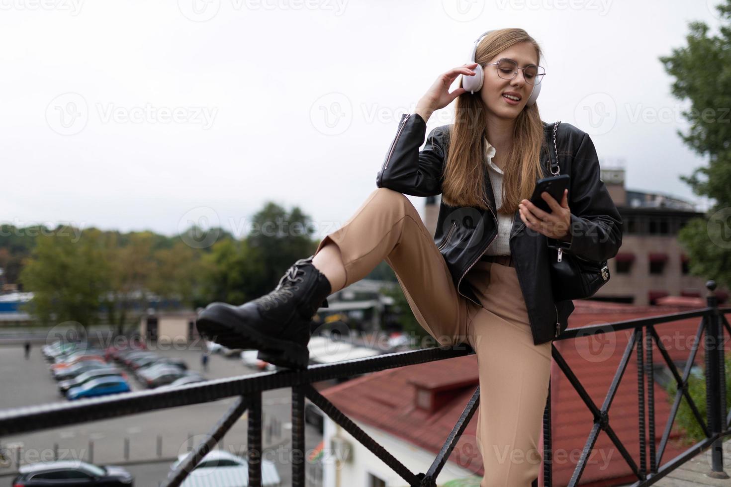 Stilvolle junge Frau in Kopfhörern hört Musik von einem Handy mit ihrem Bein auf der Brüstung auf dem Hintergrund des Stadtpanoramas foto