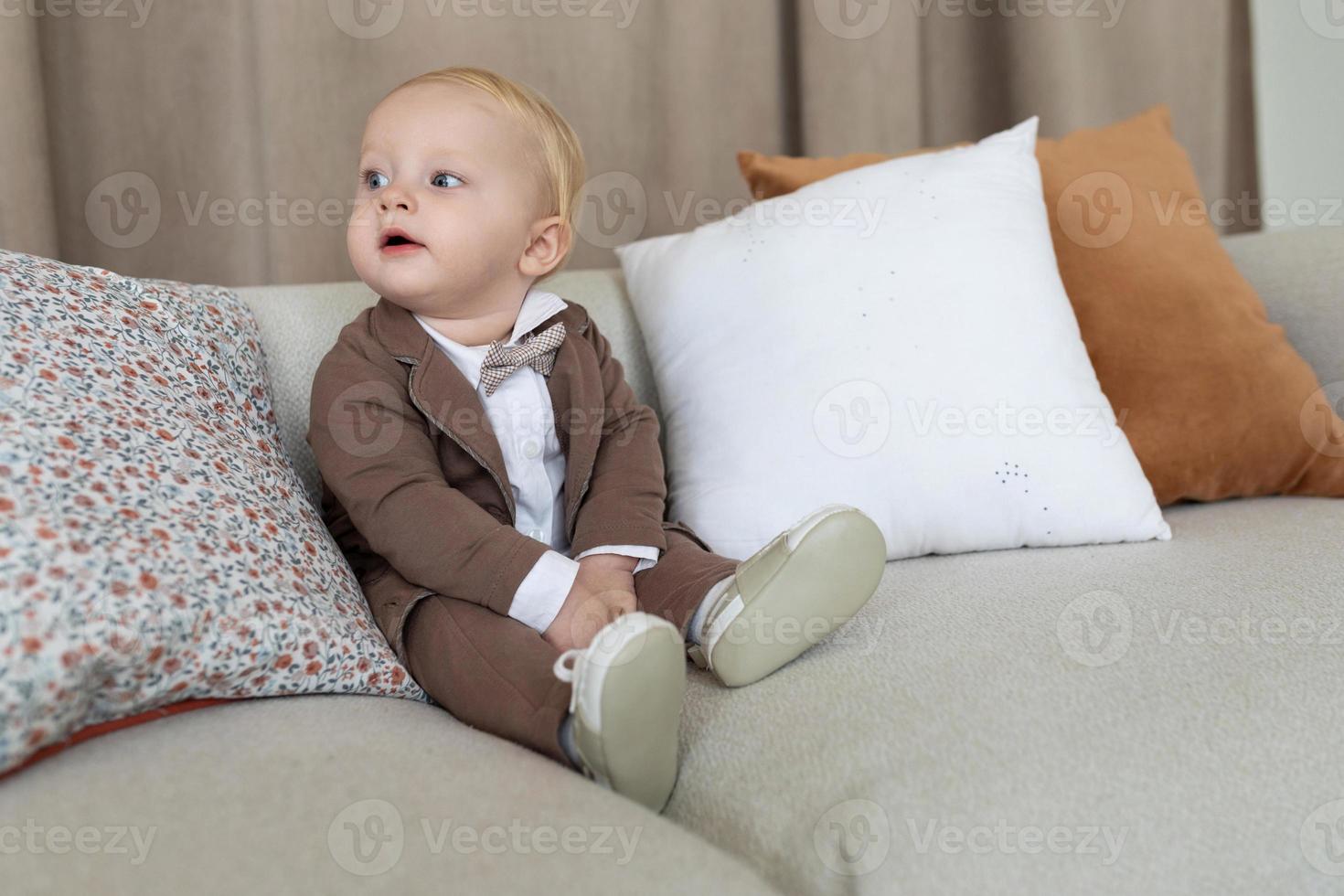 kleiner Junge in Geschäftskleidung sitzt auf der Couch und schaut sich im Zimmer um foto