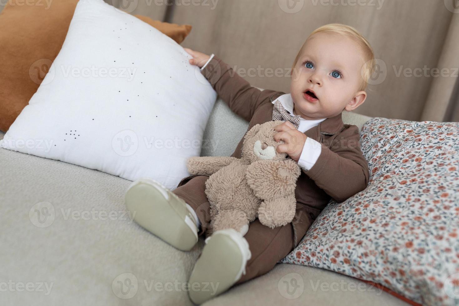 kleiner junge in stilvoller geschäftskleidung mit einem spielzeug sitzt auf einem weichen sofa foto