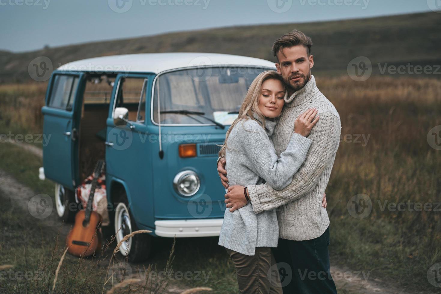 bewacht sie. schönes junges Paar, das sich umarmt und lächelt, während es in der Nähe des blauen Minivans im Retro-Stil steht foto