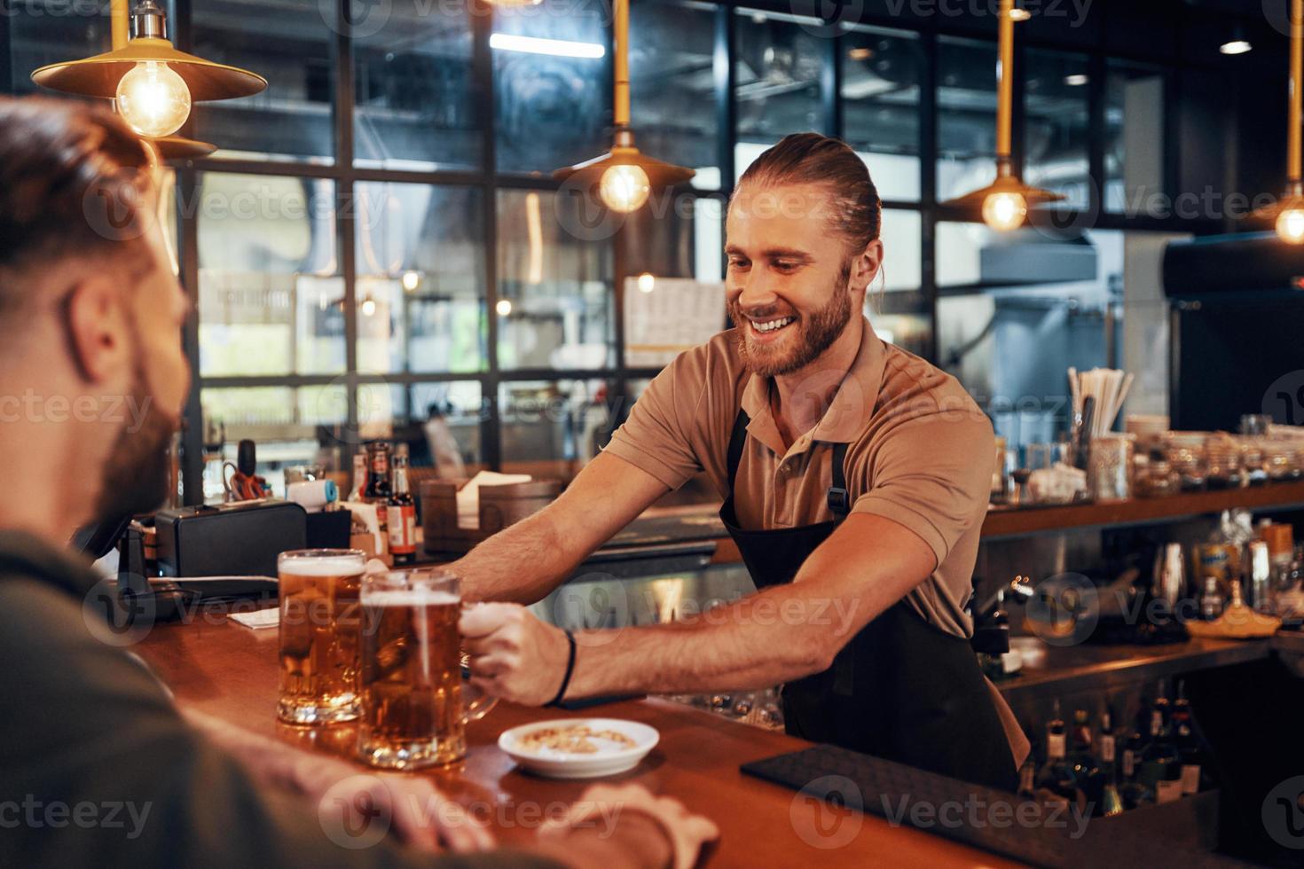 charmanter junger Barkeeper in Schürze, der Bier serviert und lächelt, während er in der Kneipe arbeitet foto