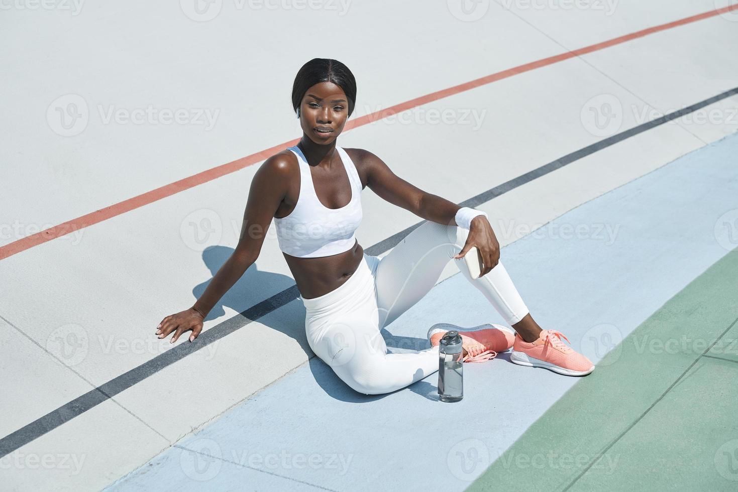 Schöne junge afrikanische Frau in Sportkleidung, die eine Flasche mit Wasser hält, während sie auf der Sportstrecke im Freien sitzt foto