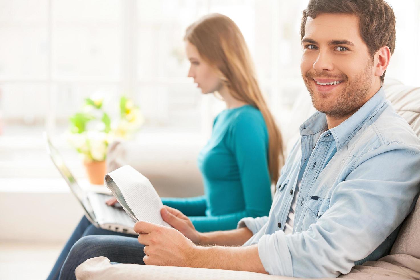 gemeinsam ihren freien Tag genießen. Schöner junger Mann, der auf der Couch sitzt und eine Zeitung hält, während seine Frau im Hintergrund sitzt und am Laptop arbeitet foto