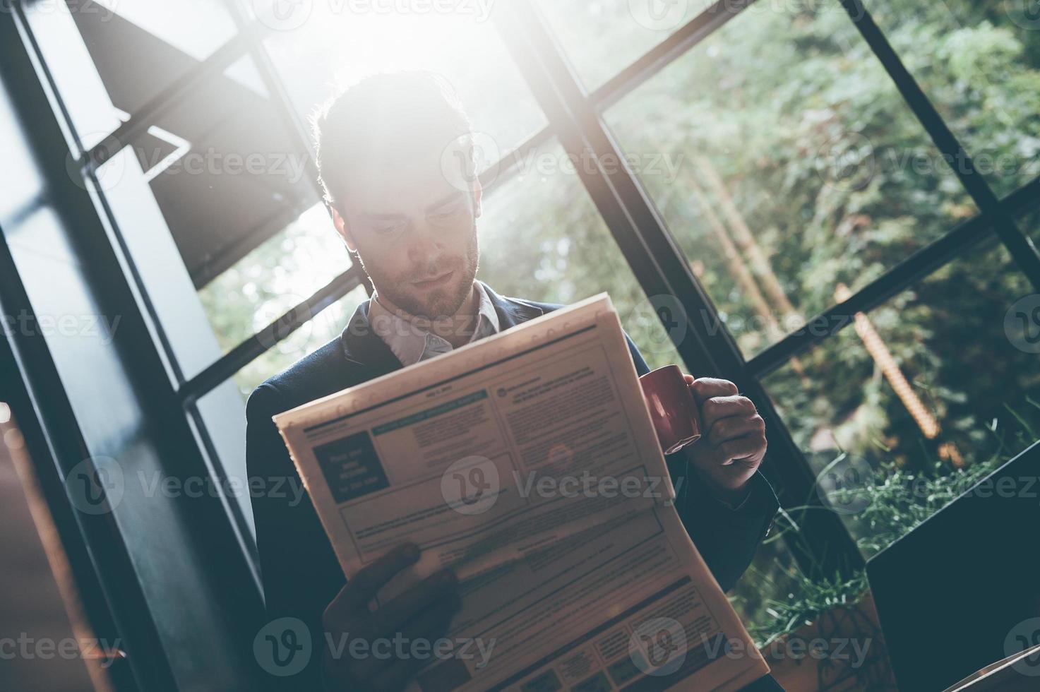 Lesen der neuesten Nachrichten. Tiefwinkelansicht eines selbstbewussten jungen Mannes, der frische Zeitung liest und eine Kaffeetasse hält, während er sich an der Fensterbank im Büro oder Café lehnt foto