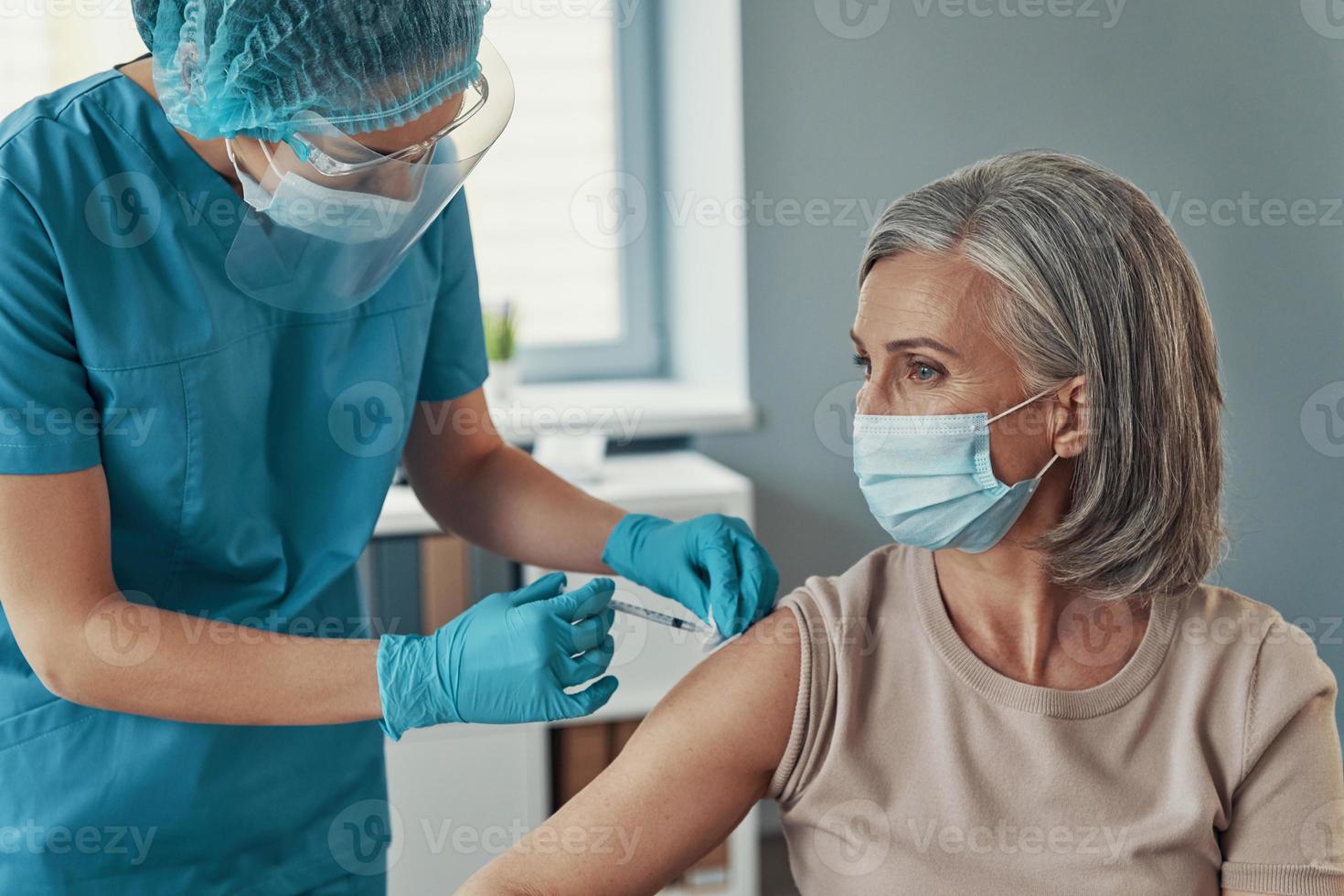 Krankenschwester in Schutzkleidung, die reife Frauen während der Arbeit im Krankenhaus impft foto