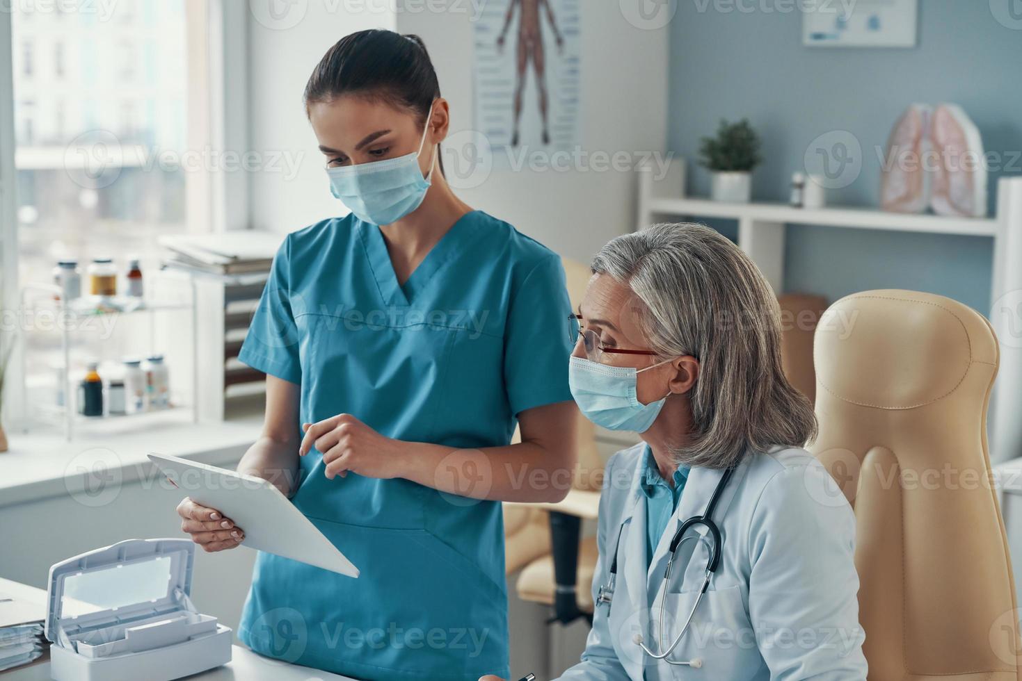 zwei mitarbeiterinnen in medizinischer uniform und schutzmasken unterhalten sich und verwenden digitale tabletten, während sie im krankenhaus arbeiten foto
