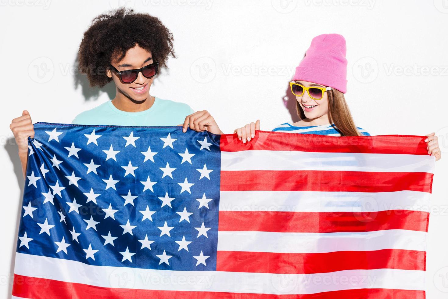 amerikanische Teenager. flippiges junges Paar, das die amerikanische Flagge vor sich hält und im Stehen vor weißem Hintergrund lächelt foto