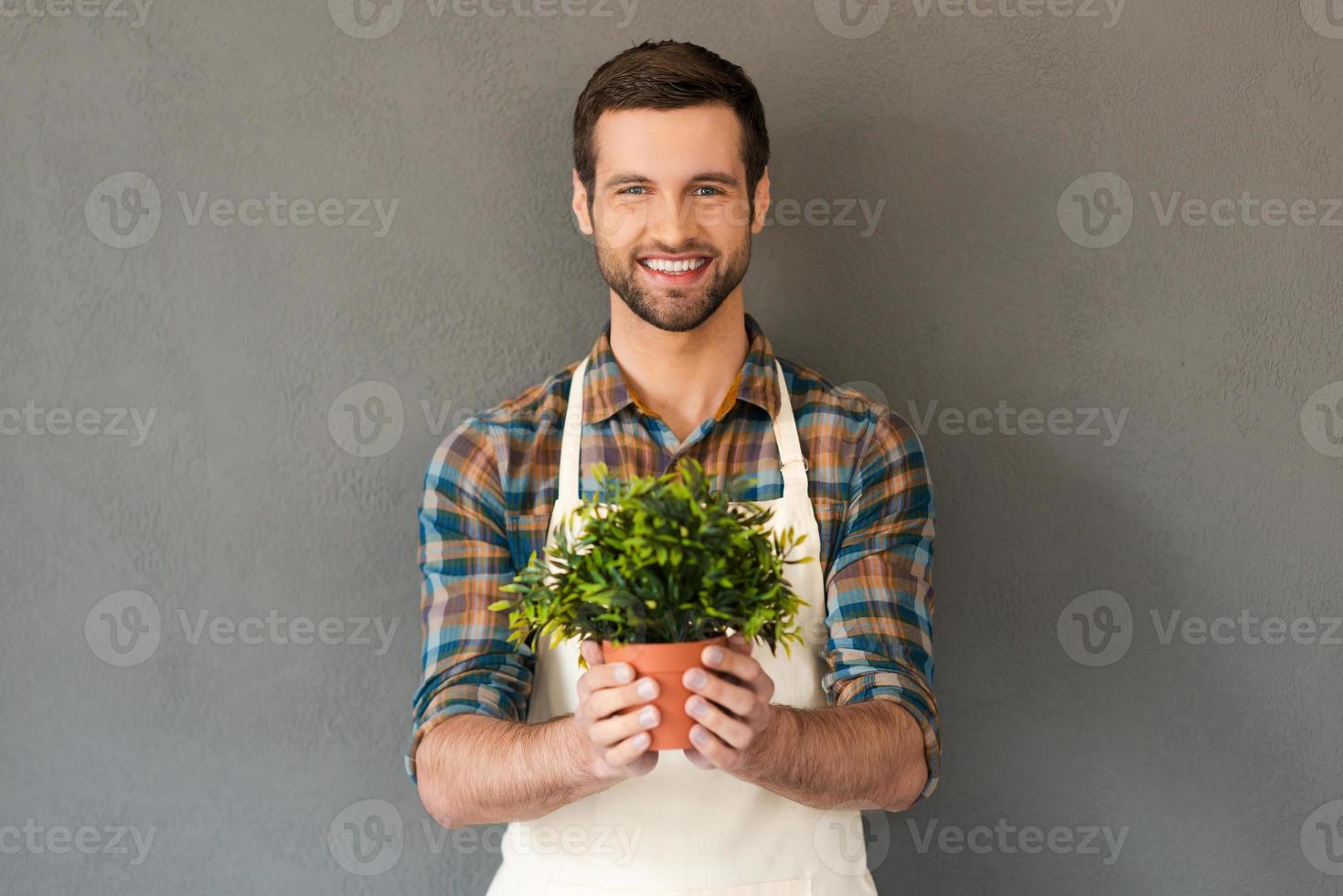 fröhlicher Gärtner. Fröhlicher junger Gärtner, der Blumentopf hält und in die Kamera lächelt, während er vor grauem Hintergrund steht foto