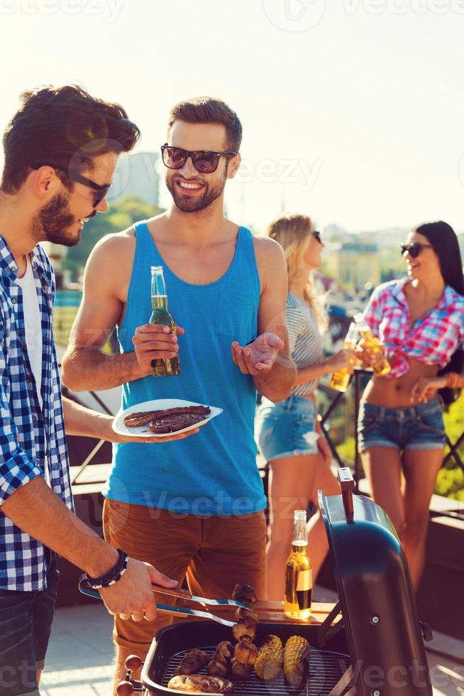 perfektes Sommermittagessen. Zwei fröhliche junge Männer grillen und lächeln, während im Hintergrund zwei Frauen miteinander reden foto