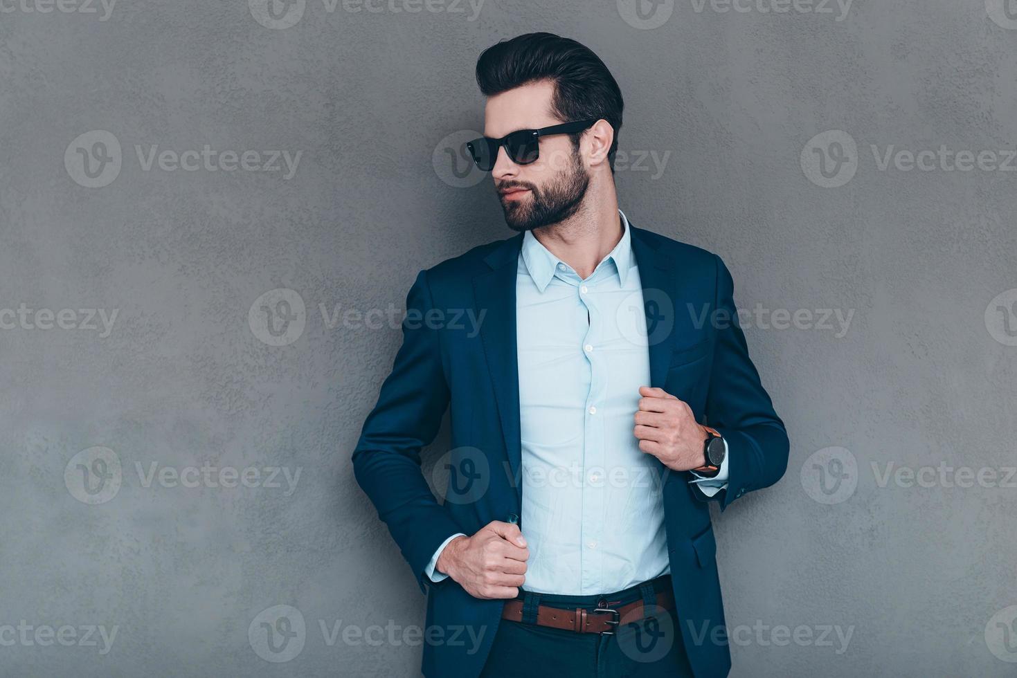 stilvoll und erfolgreich. Junger gutaussehender Mann mit Sonnenbrille, der die Hand auf seiner Jacke hält und wegschaut, während er vor grauem Hintergrund steht foto