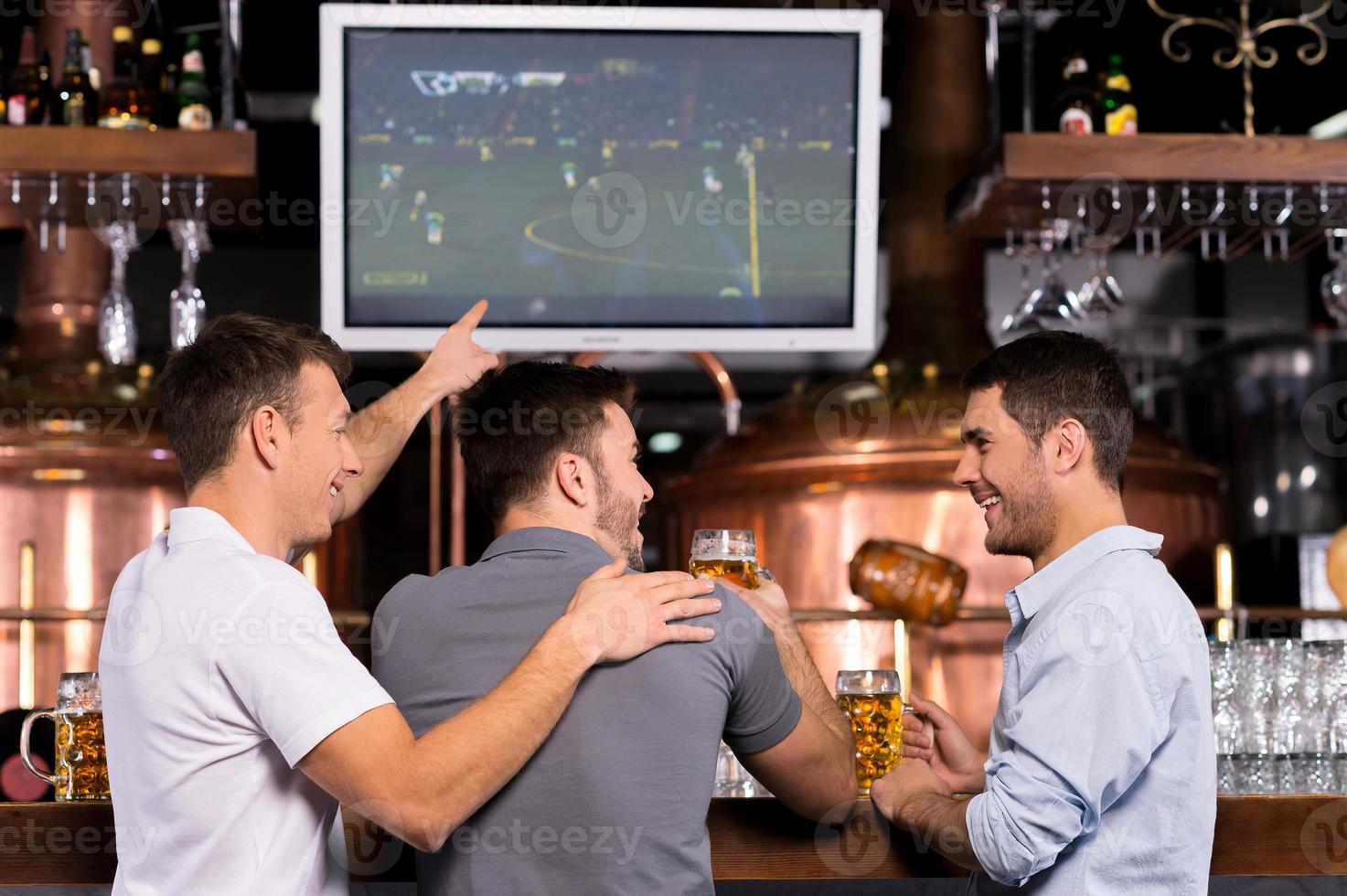 ein Fußballspiel anschauen. Rückansicht von drei glücklichen Männern, die sich ein Fußballspiel ansehen und gestikulieren, während sie in der Bar sitzen foto