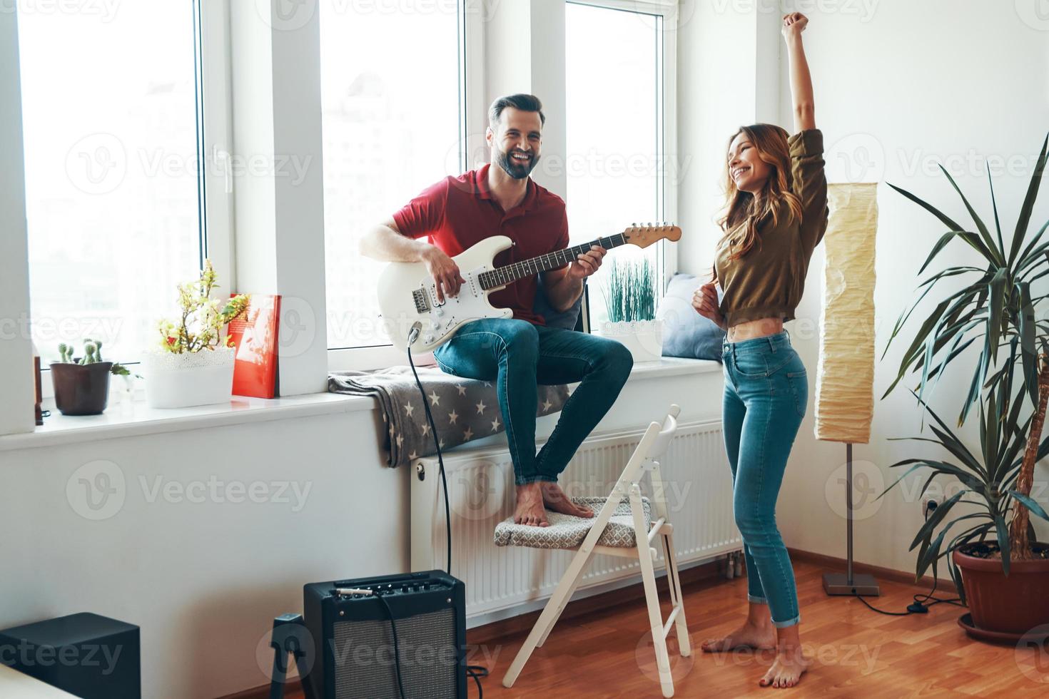 Verspieltes junges Paar in Freizeitkleidung, das sich zusammenhält und lächelt, während es Gitarre spielt foto