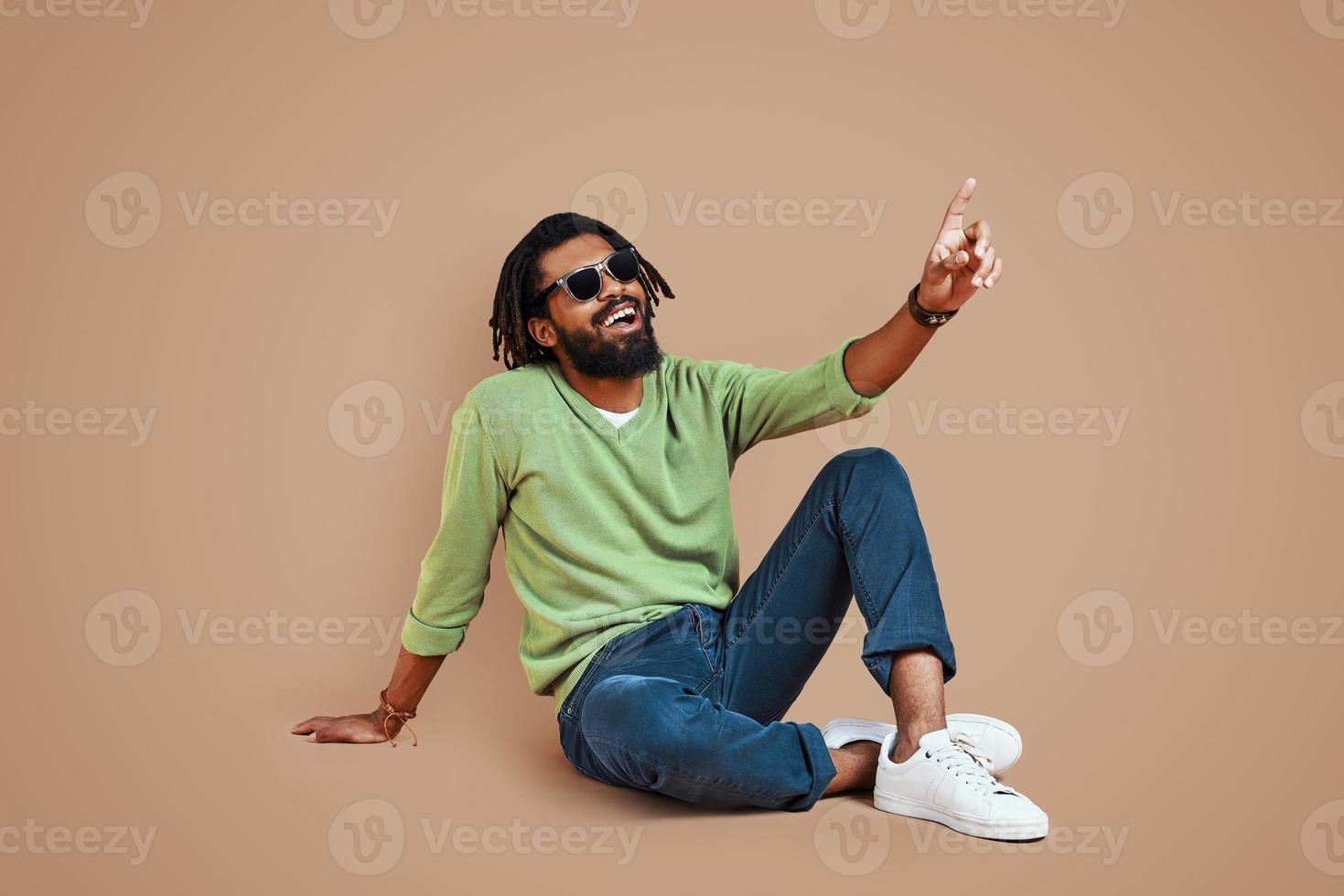hübscher junger afrikanischer mann in lässiger kleidung, der kopienraum zeigt und lächelt, während er vor braunem hintergrund sitzt foto