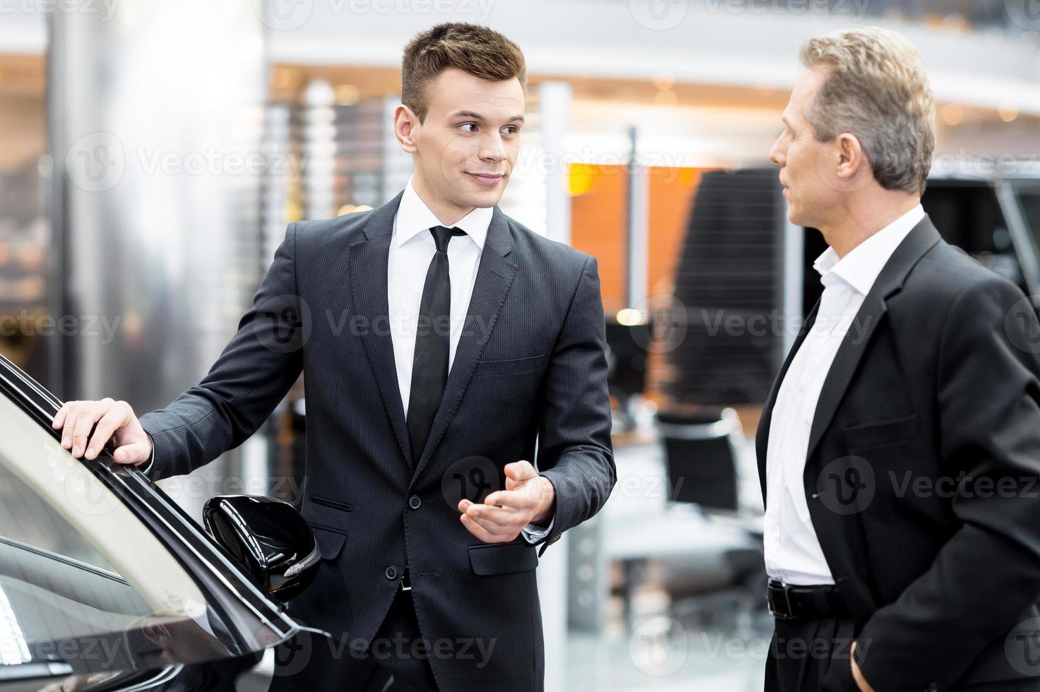 Lassen Sie mich Ihnen das Innere dieses Autos zeigen. hübscher junger klassischer autoverkäufer, der im autohaus steht und einem kunden hilft, eine entscheidung über das auto zu treffen foto