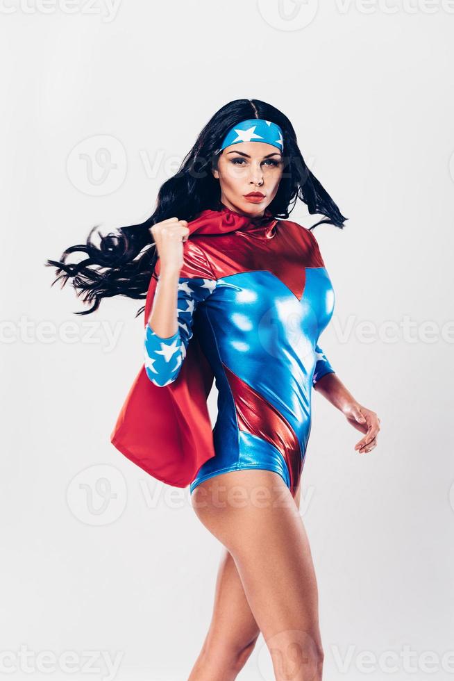Wer kann mich bekämpfen? Schöne junge Frau im Superheldenkostüm, die in die Kamera schaut, während sie in Kampfhaltung vor weißem Hintergrund steht foto