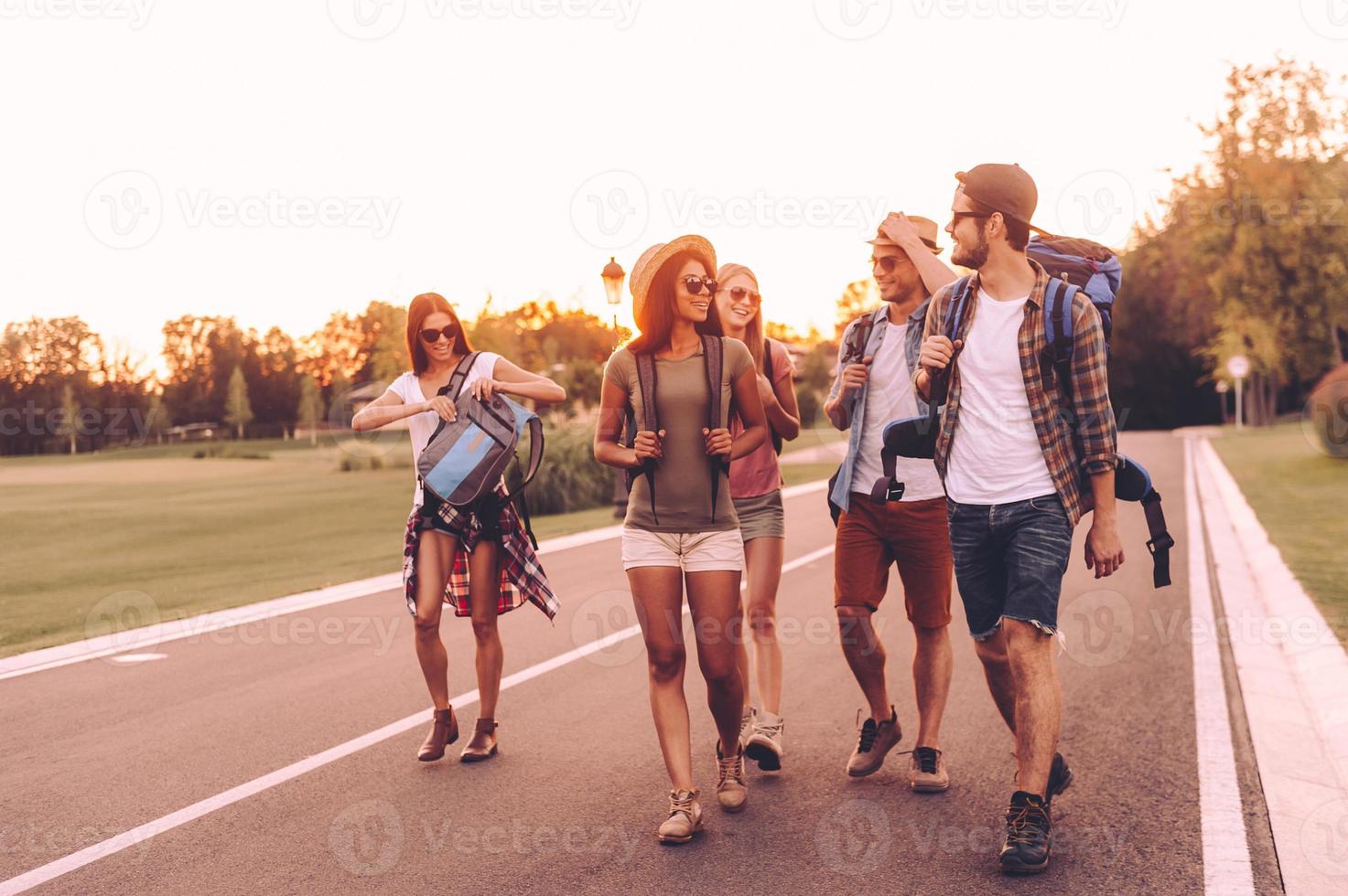 schöner tag zum wandern. Gruppe junger Leute mit Rucksäcken, die zusammen auf der Straße spazieren und glücklich aussehen foto