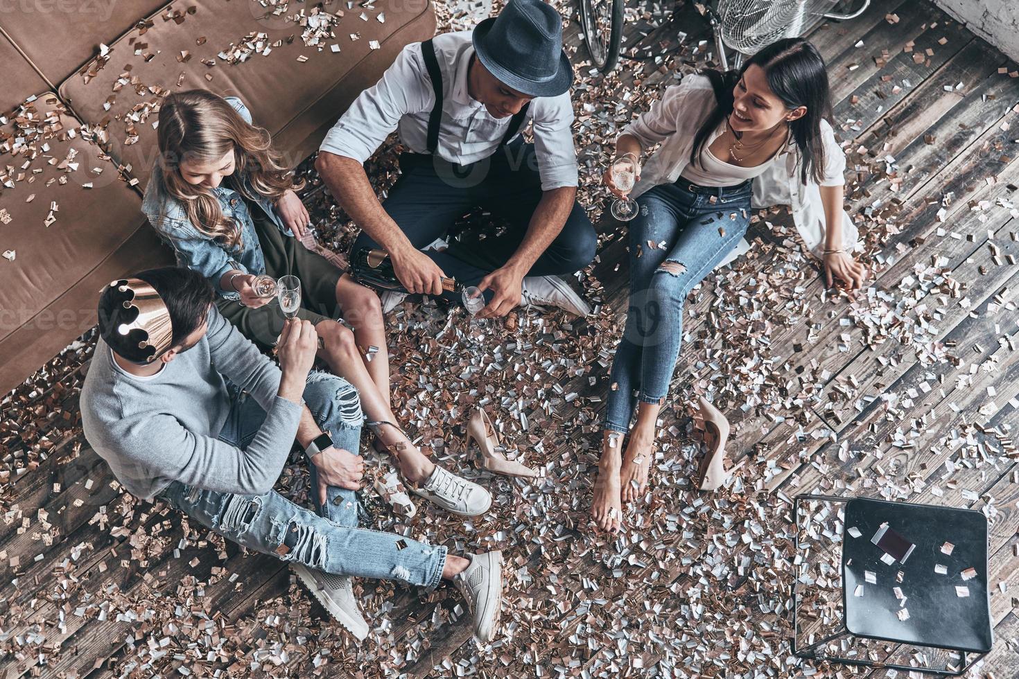 mit Freunden abhängen. Draufsicht auf moderne junge Leute, die Champagner einschenken, während sie mit Konfetti auf dem Boden sitzen foto