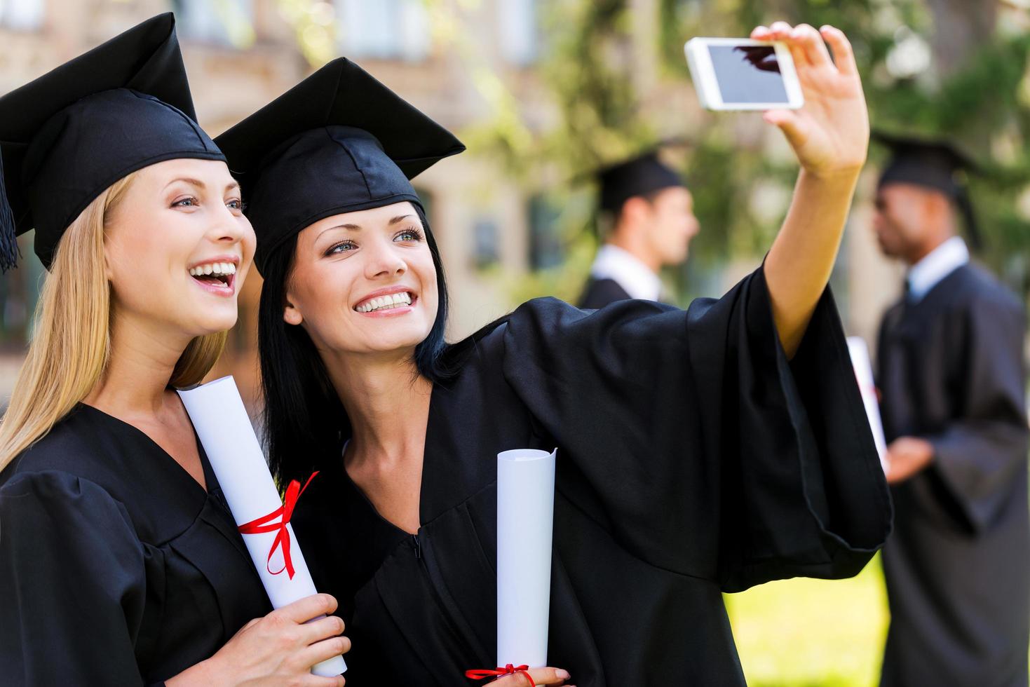 glückliche Momente festhalten. zwei glückliche frauen in abschlusskleidern, die selfie machen und lächeln, während zwei männer im hintergrund stehen foto