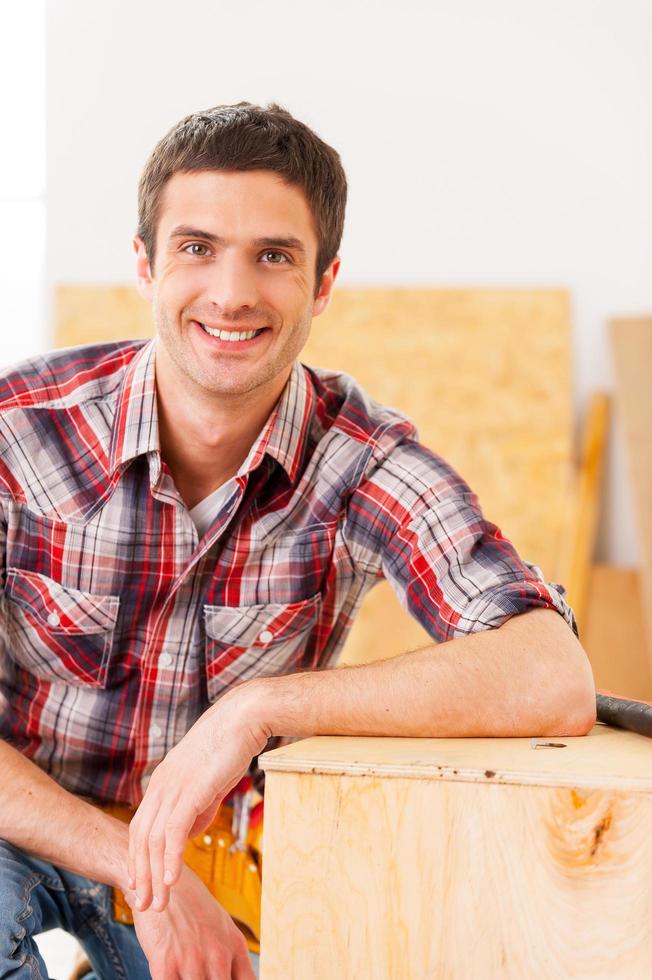 handwerker entspannt sich nach der arbeit. hübscher junger Handwerker, der in die Kamera schaut und lächelt, während er in der Werkstatt sitzt und sich an das Holzdeck lehnt foto