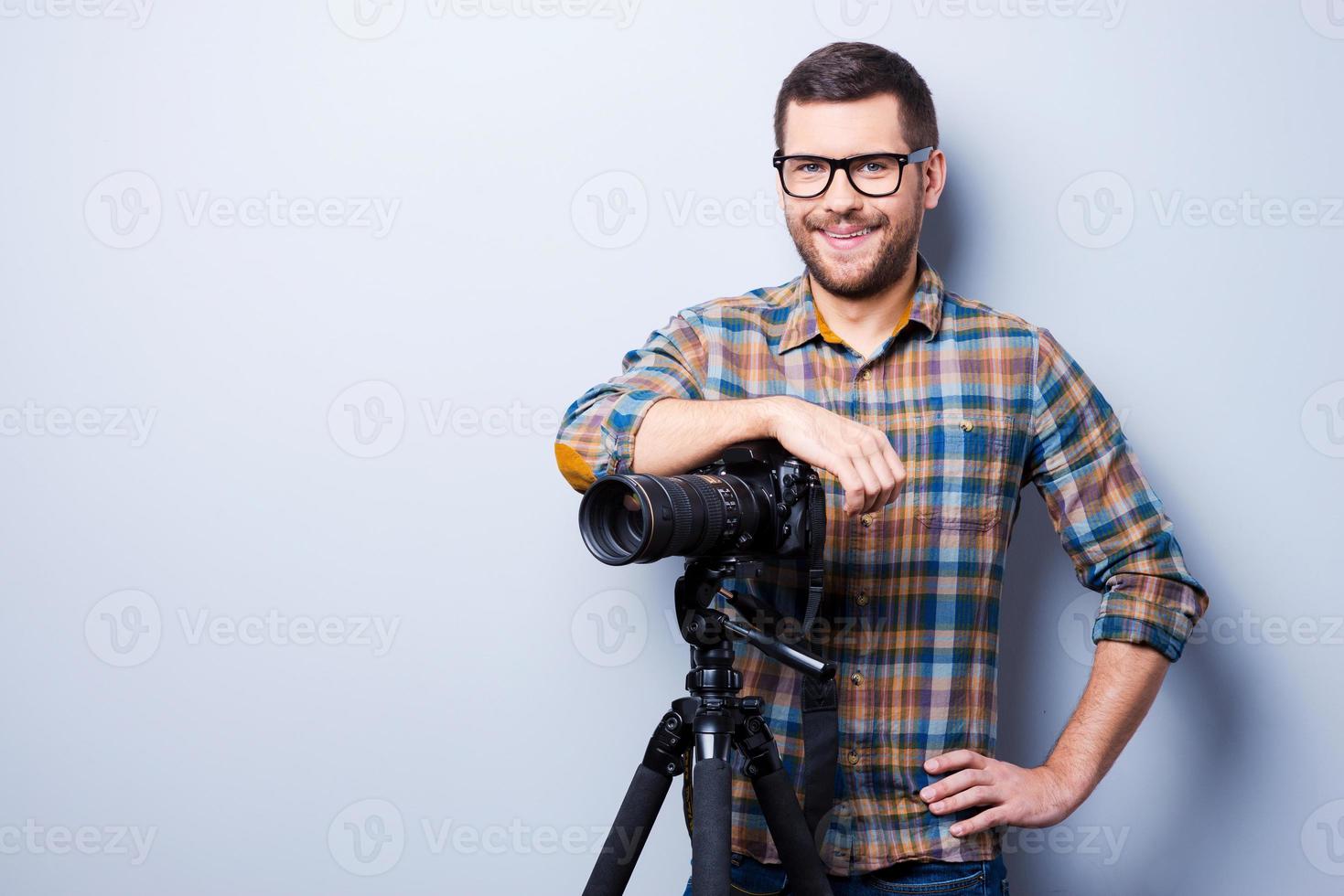 professioneller Fotograf. Porträt eines selbstbewussten jungen Mannes im Hemd, der die Hand vor der Kamera auf einem Stativ hält, während er vor grauem Hintergrund steht foto