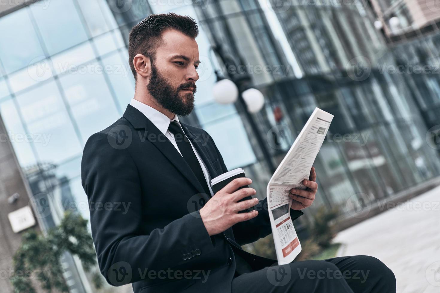 Wirtschaftsnachrichten checken. Gut aussehender junger Mann im vollen Anzug, der eine Zeitung liest, während er im Freien sitzt foto