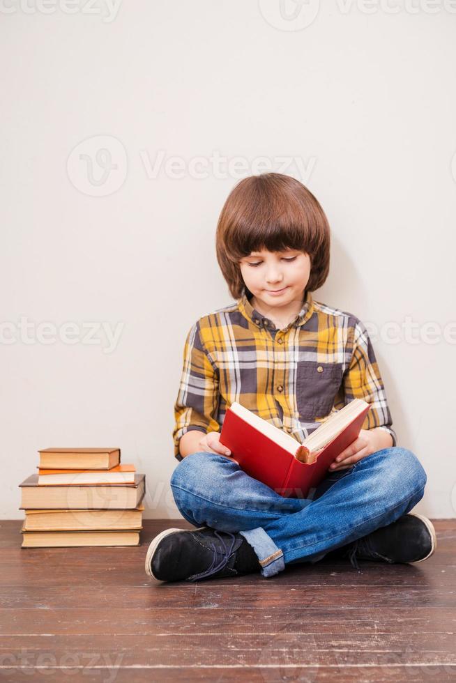 liest sein Lieblingsbuch. kleiner Junge, der ein Buch liest, während er sich an die Wand lehnt, mit einem Stapel Bücher, der neben ihm liegt foto