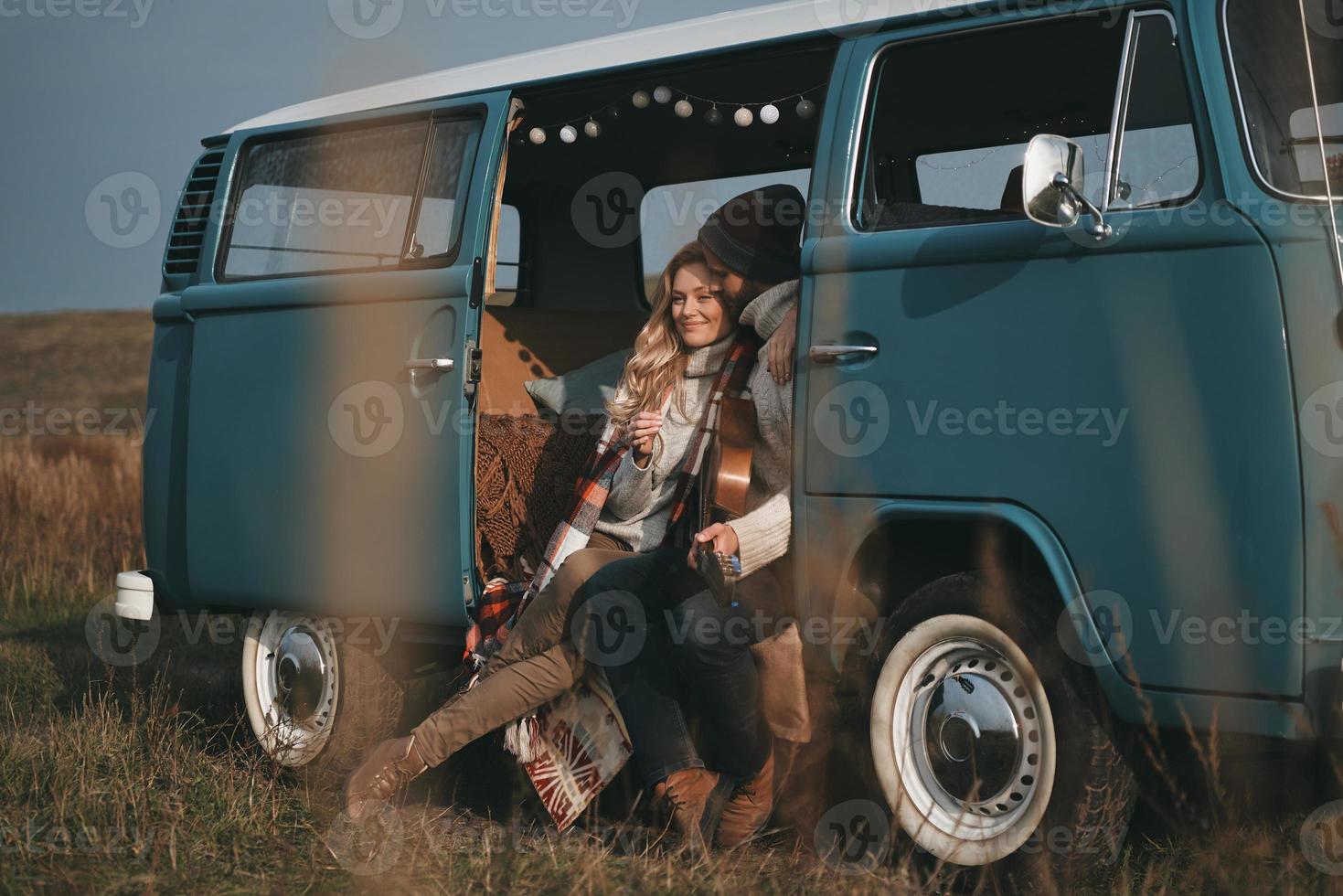 genießen ihre gemeinsame Zeit. hübscher junger Mann, der Gitarre hält und seine schöne Freundin umarmt, während er in einem blauen Minivan im Retro-Stil sitzt foto