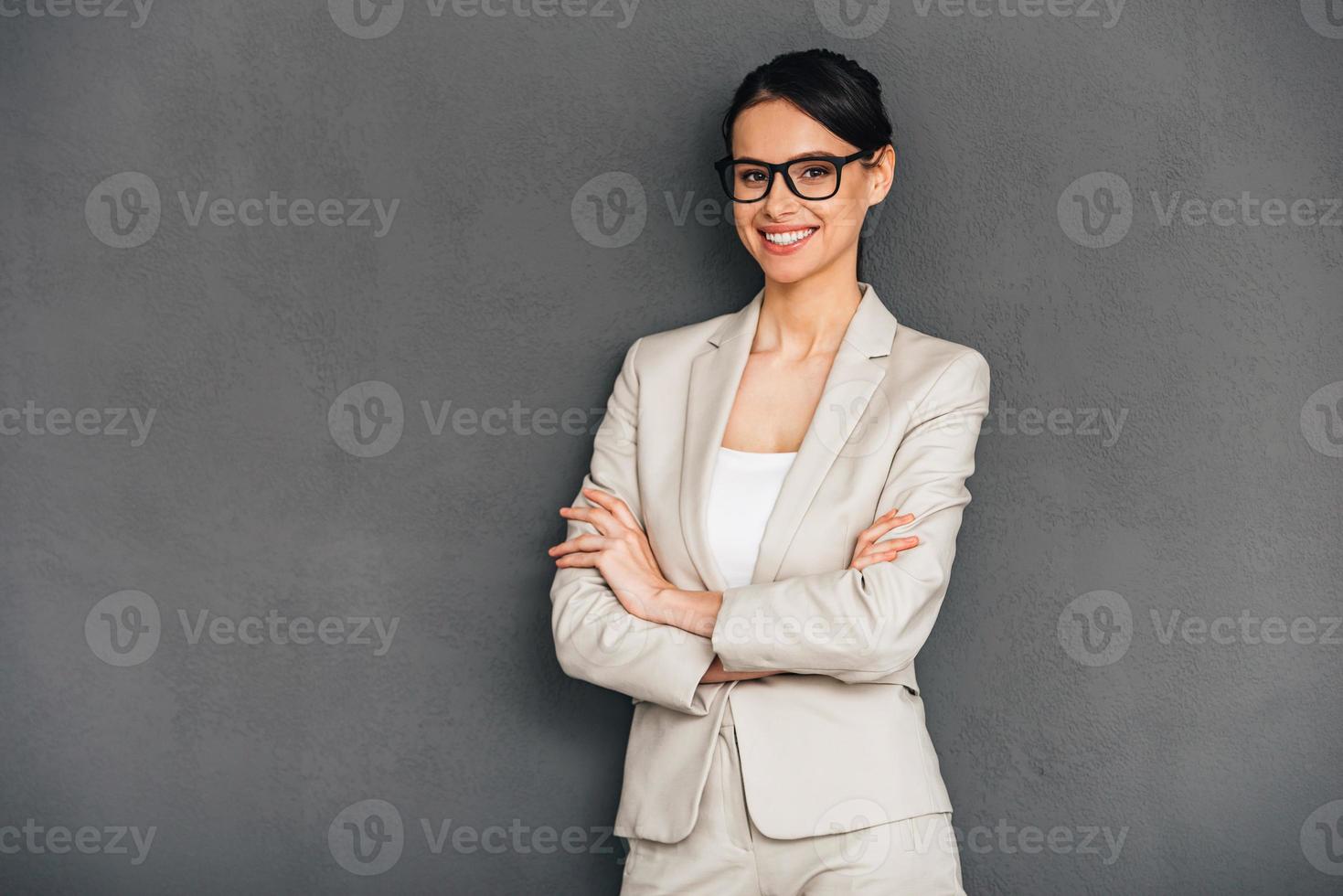 fröhlich und selbstbewusst. Fröhliche junge Geschäftsfrau mit Brille, die die Arme verschränkt hält und mit einem Lächeln in die Kamera blickt, während sie vor grauem Hintergrund steht foto