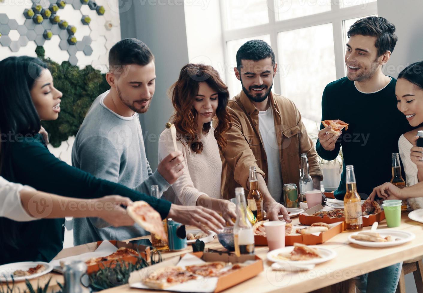 heiße Pizza genießen. Gruppe junger Leute in Freizeitkleidung, die Pizza essen und lächeln, während sie drinnen eine Dinnerparty haben foto