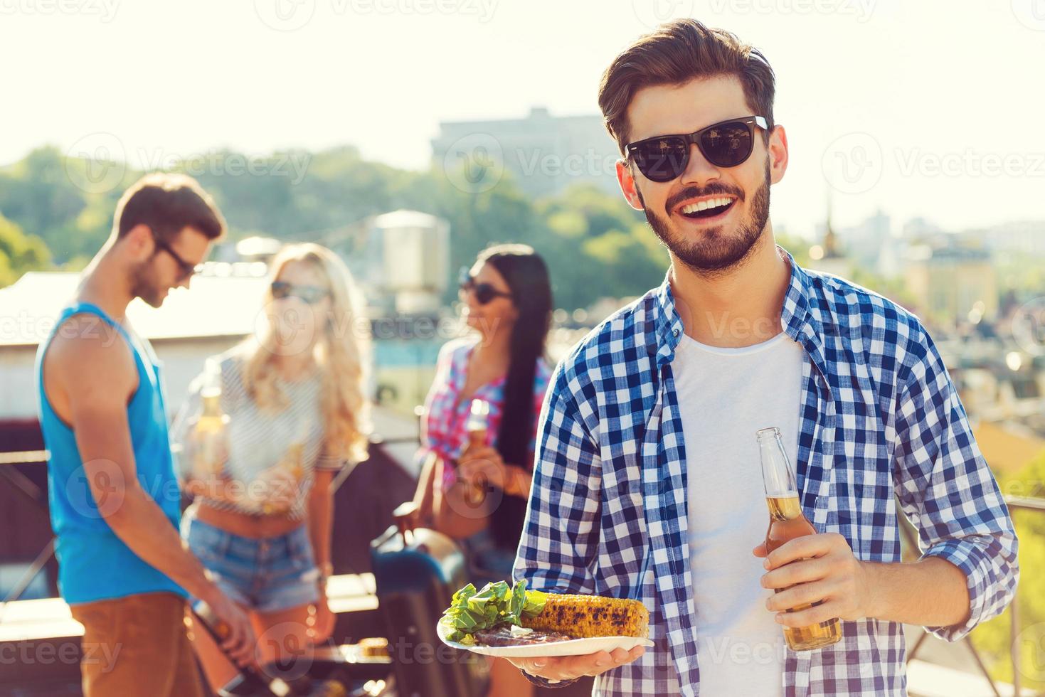 gutes essen mit besten freunden. lächelnder junger Mann, der eine Flasche mit Bier und einen Teller mit Essen hält, während drei Personen im Hintergrund grillen foto