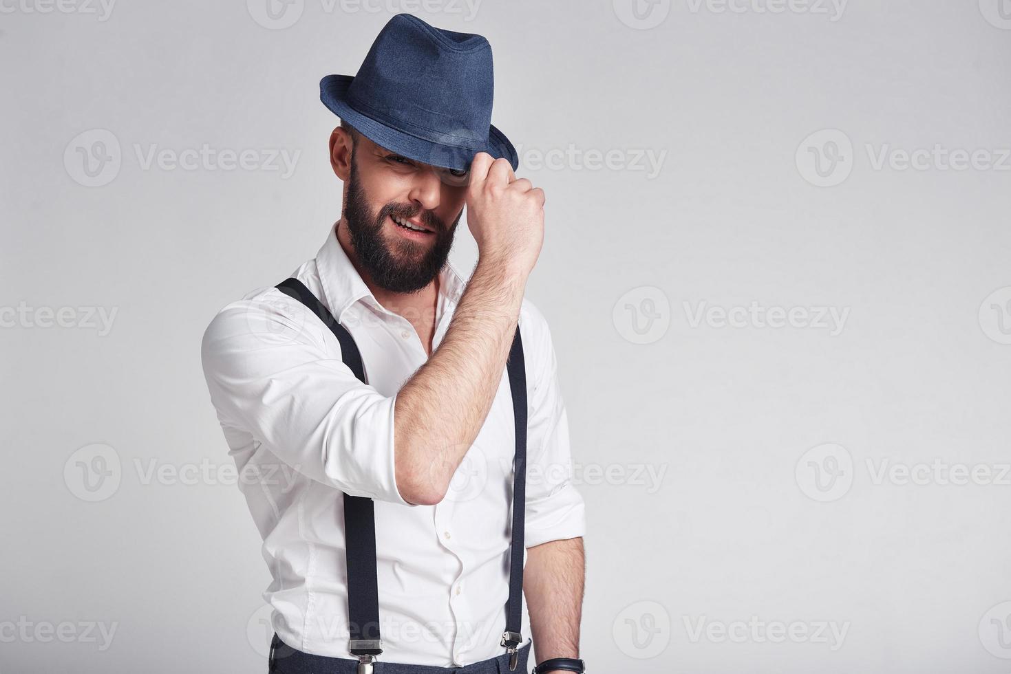 charmanter Gangster. hübscher junger Mann in Hosenträgern, der seinen Hut anpasst und mit einem Lächeln in die Kamera schaut, während er vor grauem Hintergrund steht foto
