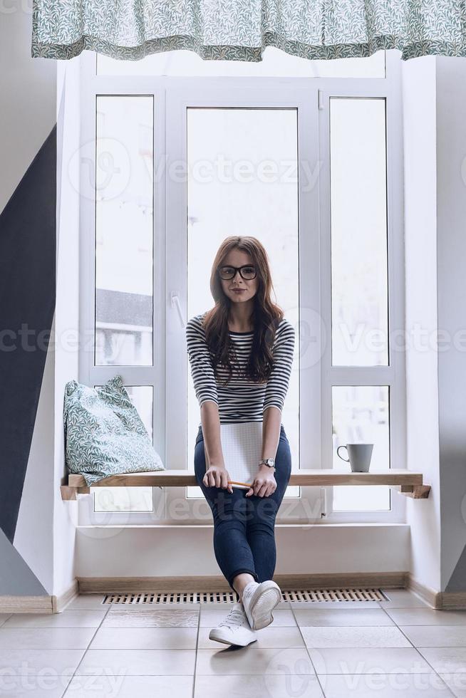 Jung und schön. attraktive junge frau, die ein notizbuch hält und in die kamera schaut, während sie auf der fensterbank sitzt foto