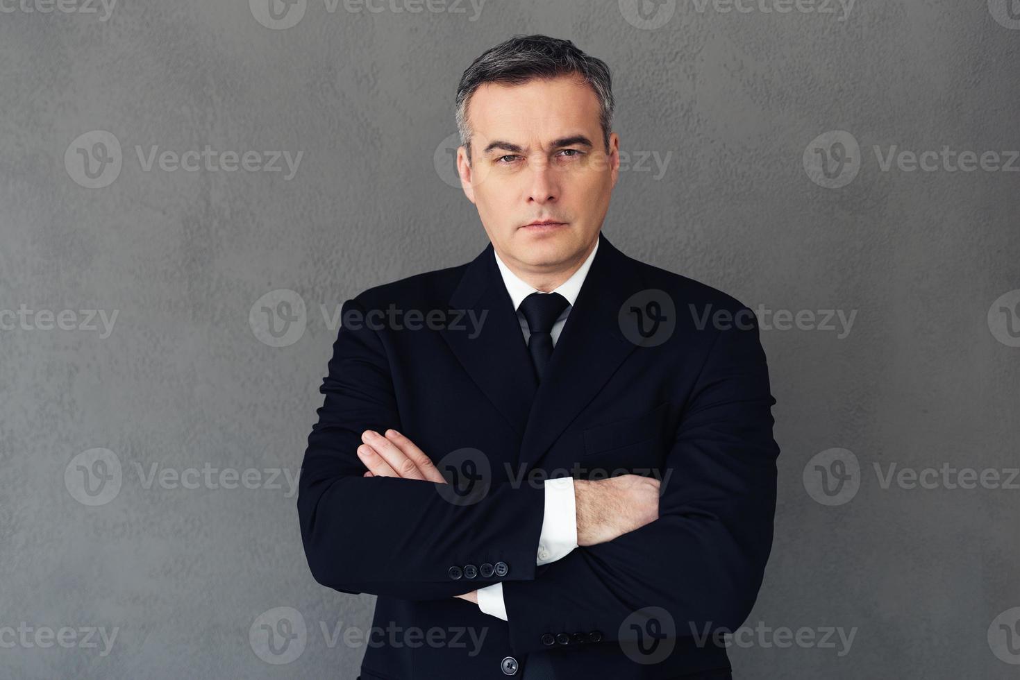 selbstbewusster Geschäftsmann. Reifer Geschäftsmann, der die Arme verschränkt hält und in die Kamera schaut, während er vor grauem Hintergrund steht foto