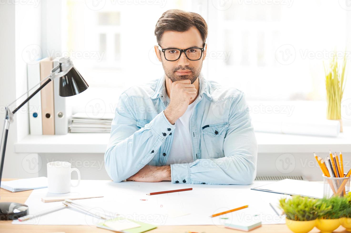 neue Ideen generieren. hübscher junger Mann in Hemd und Brille, der die Hand am Kinn hält und in die Kamera schaut, während er an seinem Arbeitsplatz sitzt foto