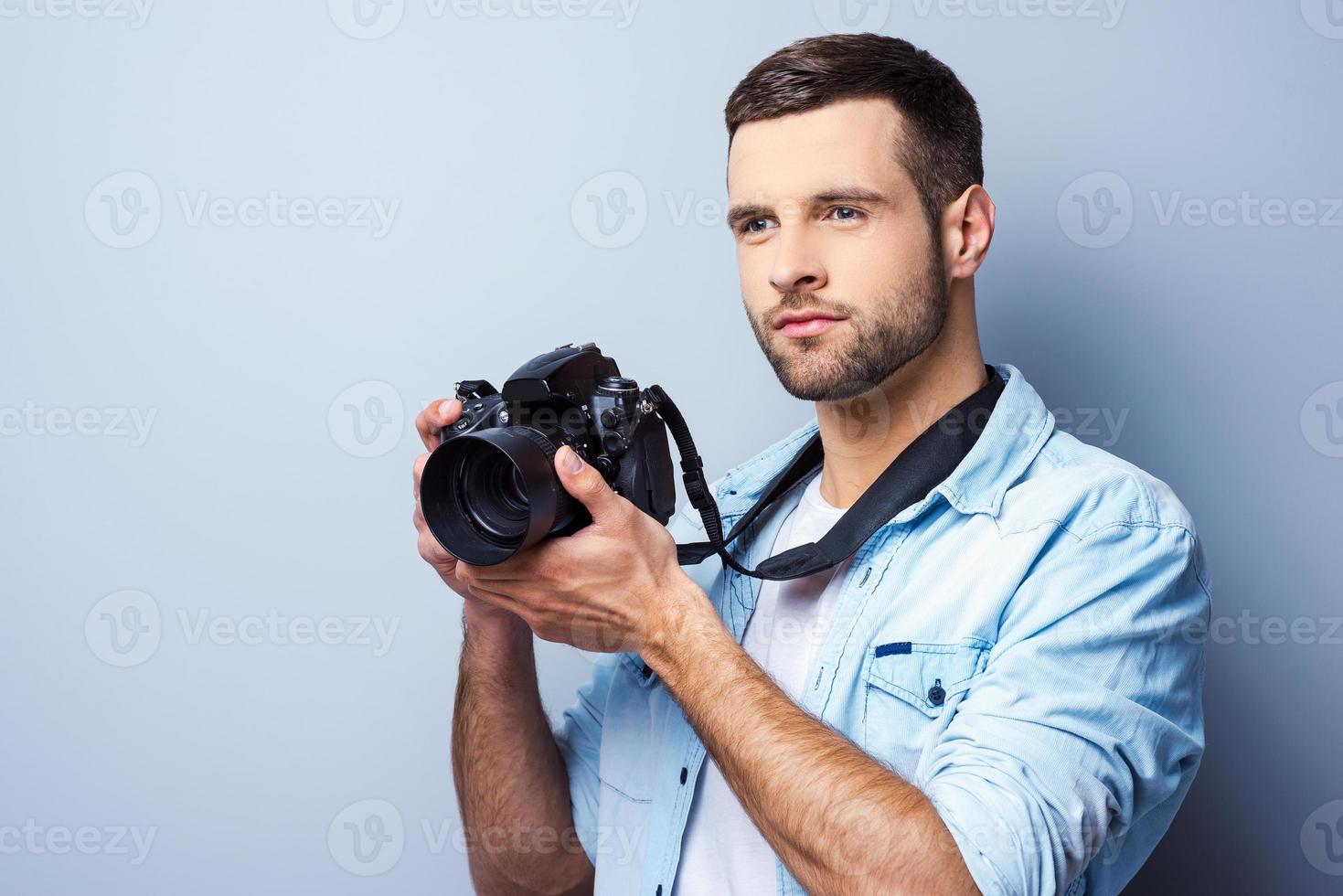 Ich werde ein großartiger Schütze sein. hübscher junger Mann, der Digitalkamera hält und wegschaut, während er vor grauem Hintergrund steht foto