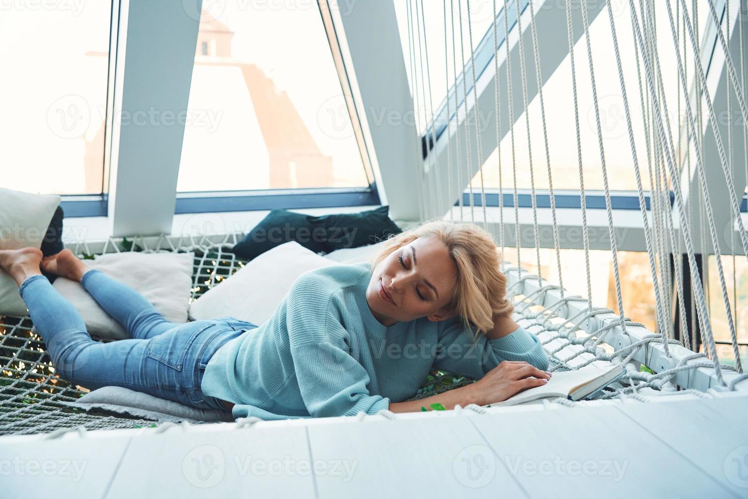 schöne junge frau, die buch liest und lächelt, während sie sich zu hause in der großen hängematte entspannt foto