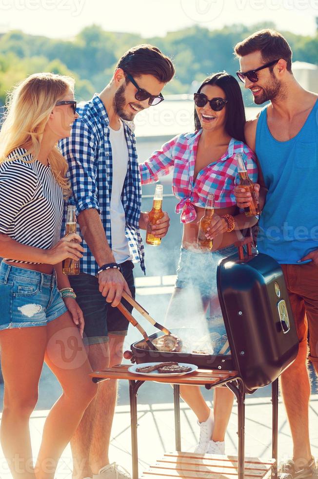 unbeschwerte Zeit mit tollen Menschen. Gruppe lächelnder junger Leute, die sich zusammenschließen und grillen, während sie auf dem Dach stehen foto