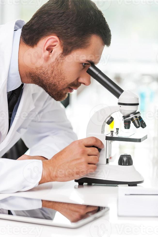 Forschung und Entwicklung. Seitenansicht eines konzentrierten jungen Wissenschaftlers in weißer Uniform mit Mikroskop, während er an seinem Arbeitsplatz sitzt foto