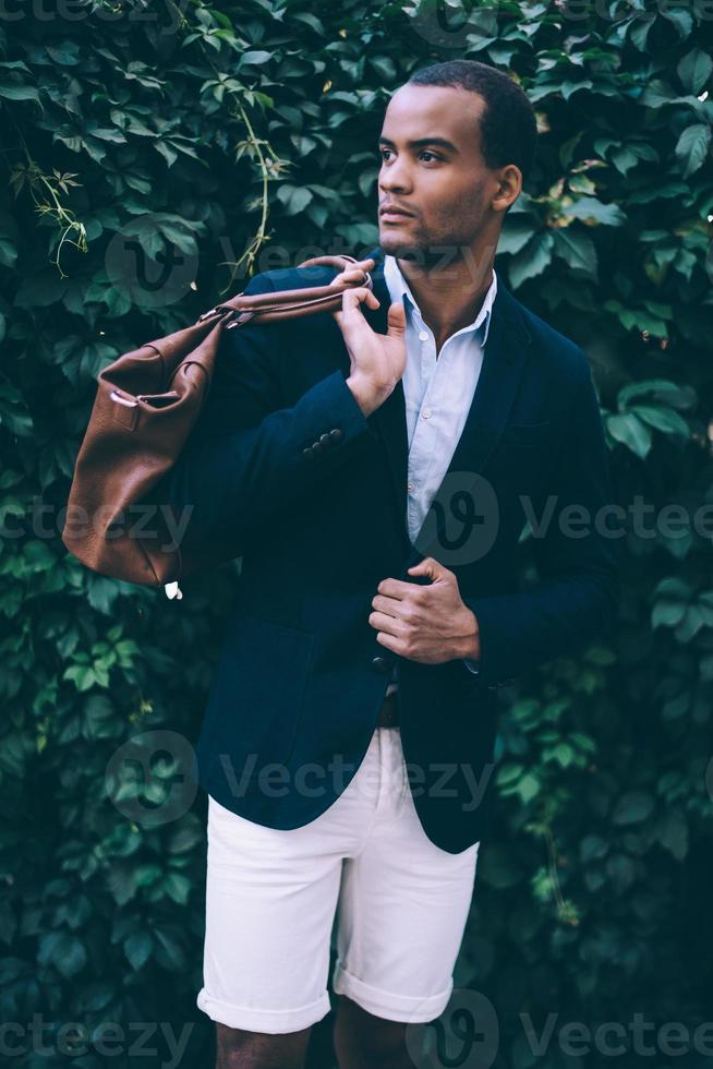 überzeugt von seinem perfekten Stil. hübscher junger afrikanischer mann in eleganter lässiger kleidung, der tasche auf der schulter trägt und wegschaut, während er im freien vor grünem pflanzenhintergrund steht foto