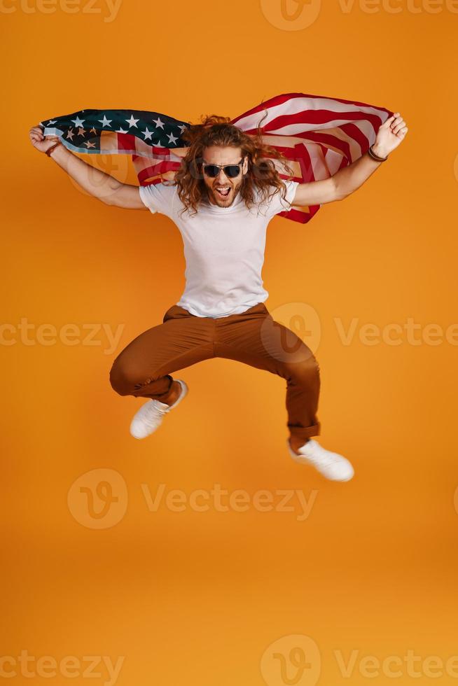 Fröhlicher junger Mann in Freizeitkleidung, der ein Gesicht macht und in die Kamera schaut, während er die amerikanische Flagge vor gelbem Hintergrund hält foto