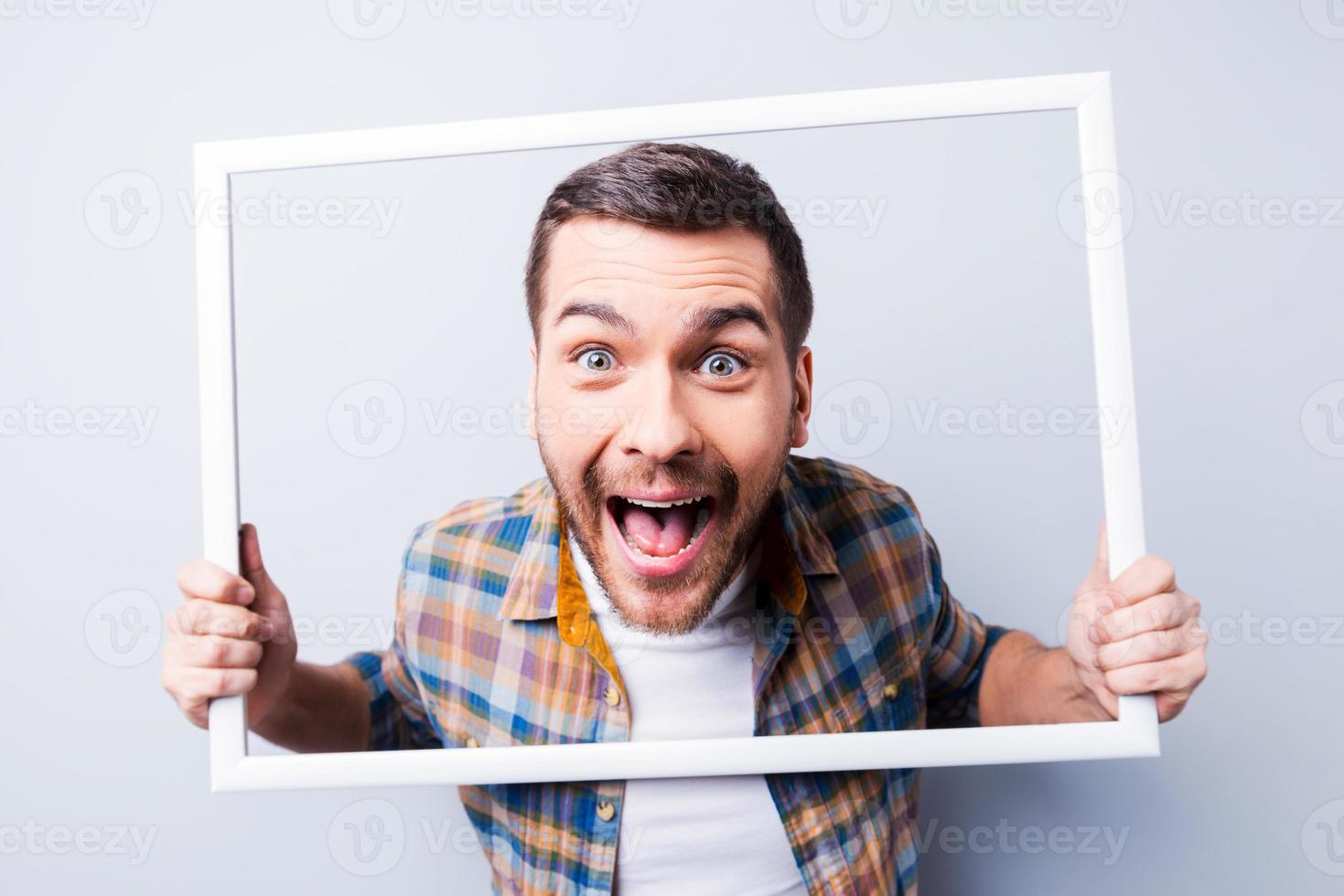 verrücktes Bild. hübscher junger Mann im Hemd, der Bilderrahmen vor seinem Gesicht hält und lächelt, während er vor grauem Hintergrund steht foto