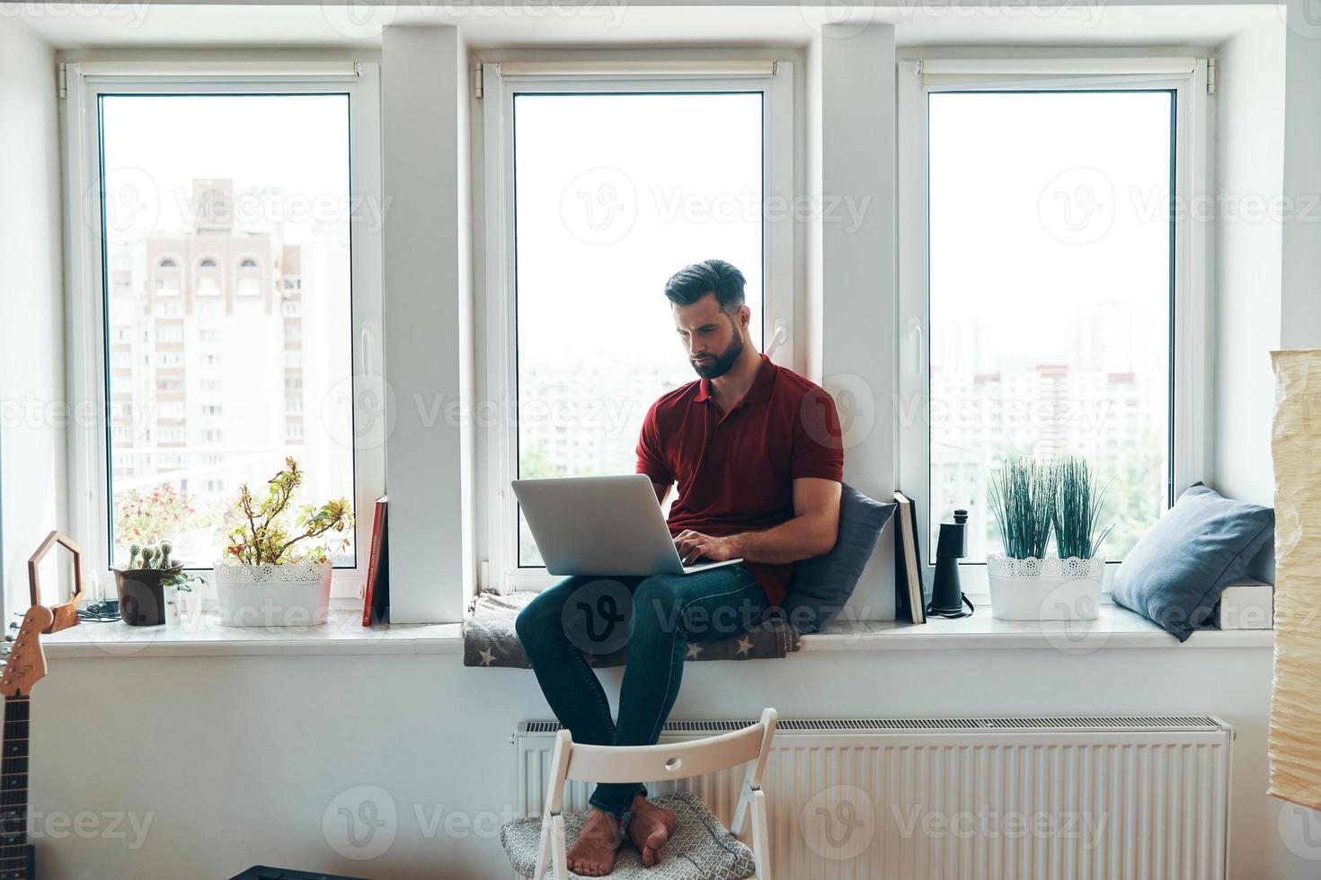 Beschäftigter junger Mann in Freizeitkleidung mit Laptop, während er auf der Fensterbank sitzt foto