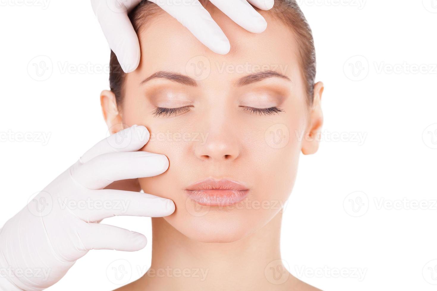 Vorbereitung vor Gesichtsoperationen. Schöne junge Frau, die die Augen geschlossen hält, während der Arzt in medizinischen Handschuhen ihr Gesicht isoliert auf Weiß untersucht foto