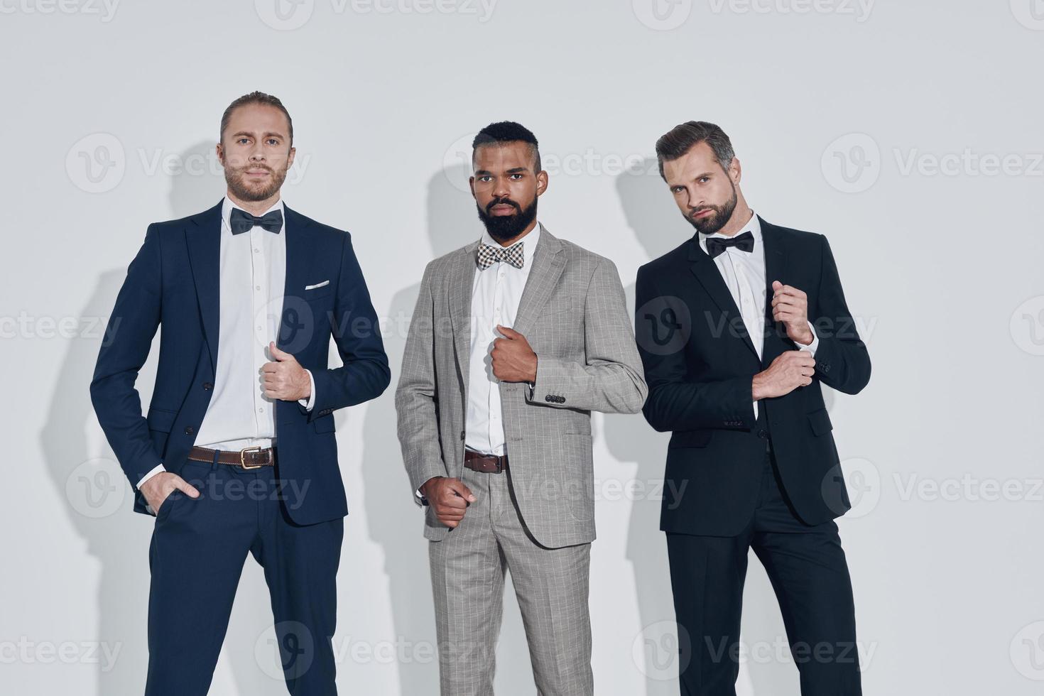 Drei hübsche junge Männer in Anzügen und Schleifen, die in die Kamera blicken, während sie vor grauem Hintergrund stehen foto