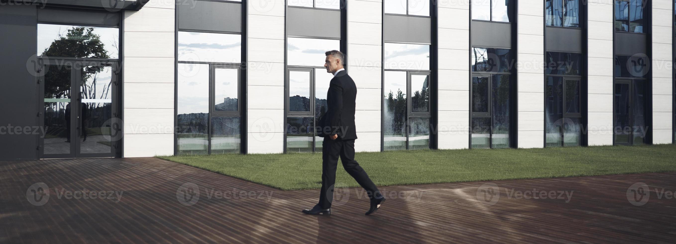 Selbstbewusster, reifer Geschäftsmann, der vor dem Bürogebäude spazieren geht foto
