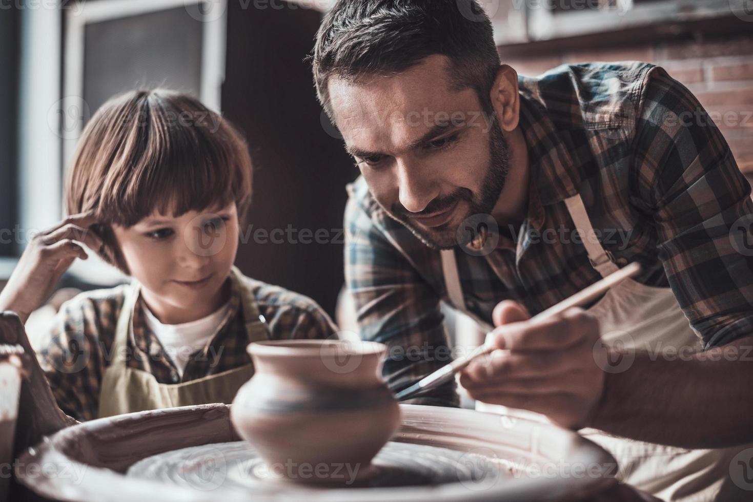 Er lernt gerne neue Fähigkeiten. kleiner Junge, der in der Töpferklasse einen selbstbewussten jungen Mann anschaut, der auf Keramiktopf zeichnet foto