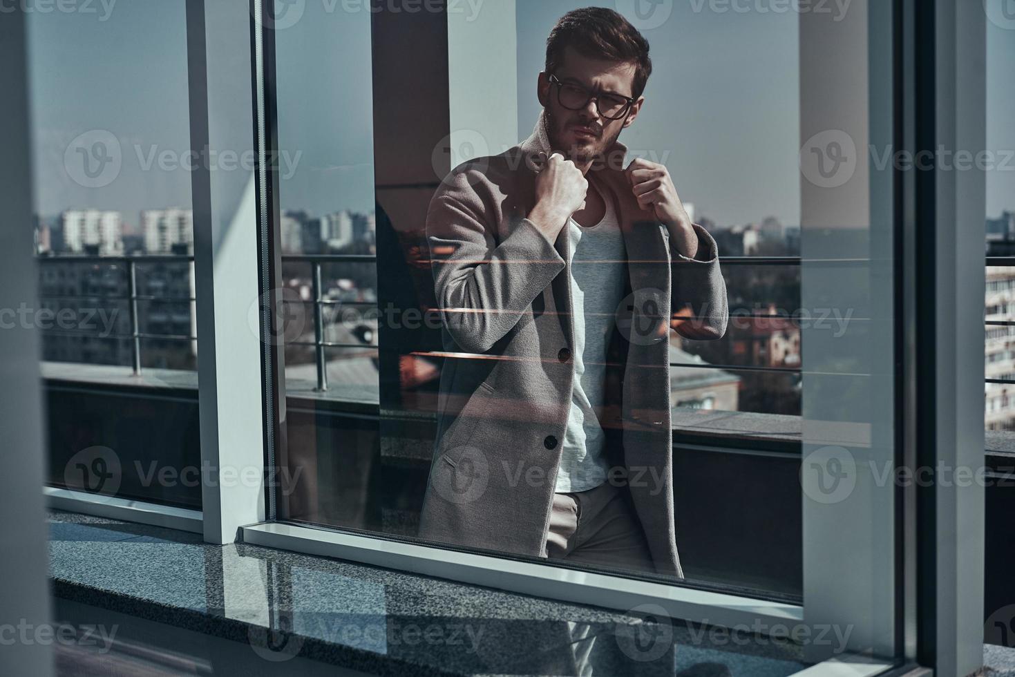 perfekter Stil. durch das fenster schoss ein hübscher junger mann in brillen, der seine jacke anpasst, während er im freien geht foto