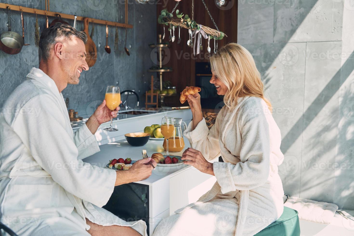 Schönes reifes Paar in Bademänteln, das zusammen frühstückt, während es Zeit in der heimischen Küche verbringt foto