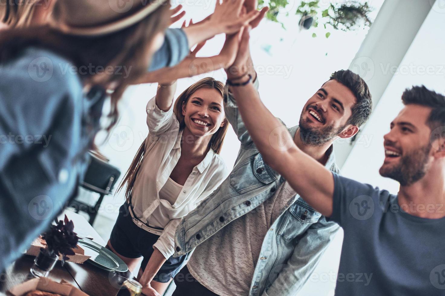 enge Freunde. Gruppe junger Leute, die sich als Symbol der Einheit High-Five geben und lächeln, während sie drinnen eine Dinnerparty haben foto