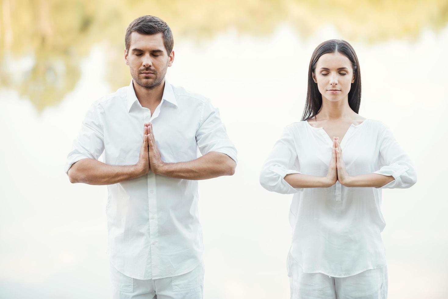 Ruhe finden. Schönes junges Paar in weißer Kleidung, das gemeinsam im Freien meditiert und die Augen geschlossen hält foto